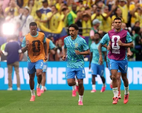 Brazil players in turquoise patterned shirts jog on a pitch during warm-up, with a crowd in the background