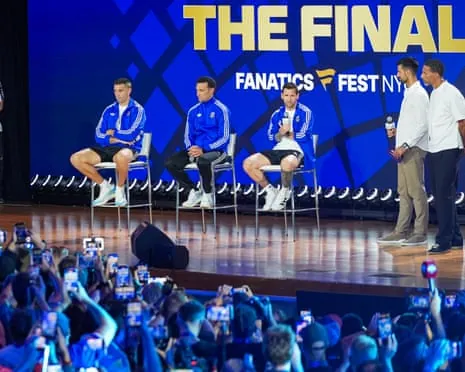 Emiliano Martinez, Lionel Scaloni and Lionel Messi sit on chairs on a stage while two others stand nearby, facing a crowd