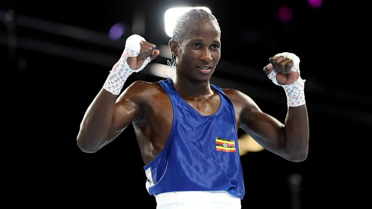 Getty Images Nuhu Batte, a boxer wearing a blue top with a small Ugandan flag printed on it, holds his fists up in the air while standing in a boxing ring.